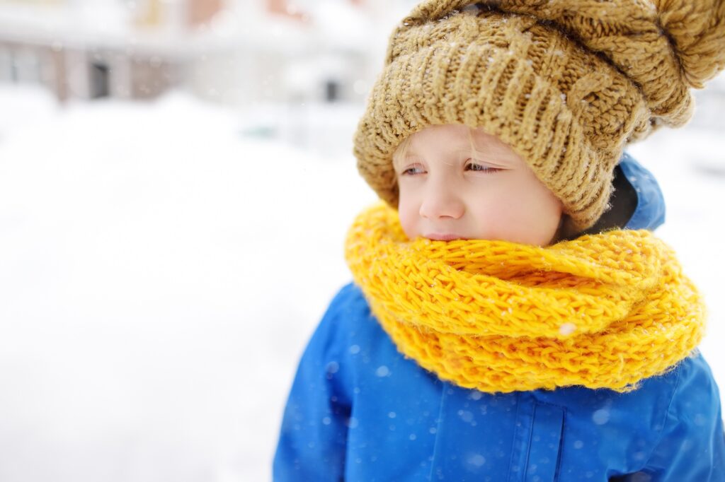 Upset little boy in blue winter clothes walks during a snowfall.